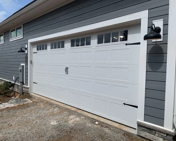A white carriage house garage door with black strap hinges and handles on a new home with dark grey lap siding